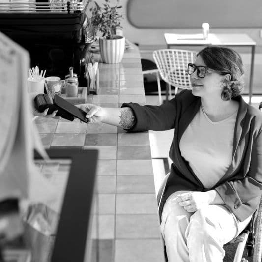 A lady who is a wheelchair user tapping her phone on a EFTPOS machine at a cafe